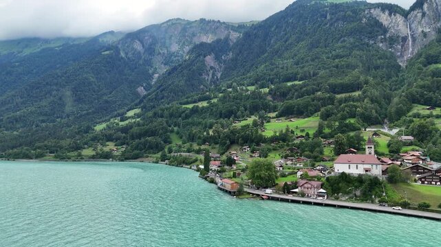Drone flies forward over turquoise Lake Brienz revealing charming Swiss alpine village of Brienz with church, chalets, and dramatic misty mountains.