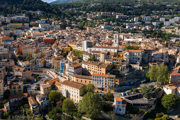 Fototapeta premium Aerial view of Grasse, France, shows terracotta rooftops, pastel facades, and the Cathedral Notre Dame du Puy above winding streets, forested hills, and modern blocks.