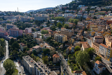 Fototapeta premium Aerial view of Grasse, France, with terraced hills, pastel buildings, red tiled roofs, winding streets, a church spire, and a curving boulevard in late afternoon light.