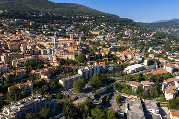 Fototapeta premium Aerial view of Grasse, France shows terracotta roofs on slopes, church towers, a roundabout, cypress and plane trees, and Alpine foothills in clear morning light.