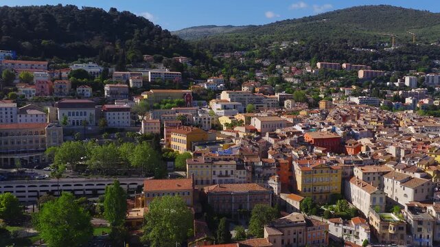 Aerial high angle pan shows Grasse, France old town, terracotta roofs, pastel facades, and a stone bell tower, with wooded hills and villas under clear midday light.
