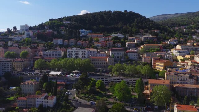 Aerial view of Grasse, France shows pastel tiers, terracotta roofs, promenade, parking terrace, white tents, flags, and sweeping motion in late morning light.