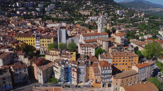 Aerial view of Grasse, Provence, France shows the bell tower cathedral and a cylindrical medieval tower, pastel facades, winding streets, cars, pedestrians, and scaffolding.