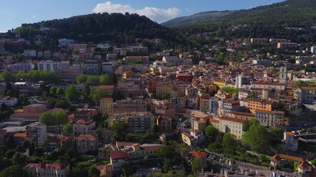 Aerial pan shows Grasse, France, hillside roofs, bell tower, winding streets, mixed era blocks, tree lined boulevards, midday light, wide panoramic view.