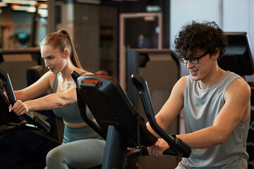 Fototapeta premium Young adult Caucasian woman and man exercising on stationary bikes in gym, supporting fitness routine and active lifestyle. Useful for wellness marketing, training ads, health campaigns