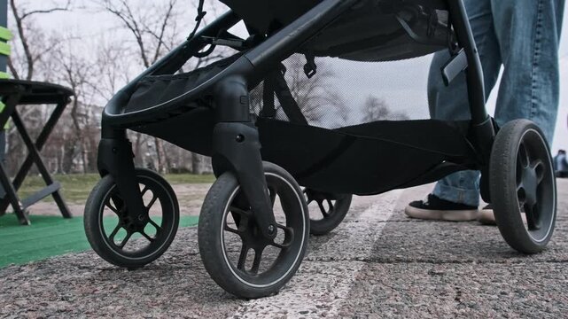 A low angle close-up shows baby stroller wheels moving smoothly on asphalt. The ground-level perspective highlights the mechanics of the wheels and the urban road texture.