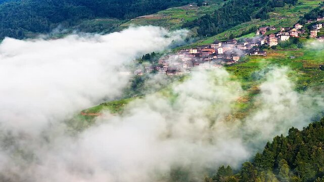 Aerial view of traditional rural village built on green terraced hillsides under moving clouds in Guilin, China.