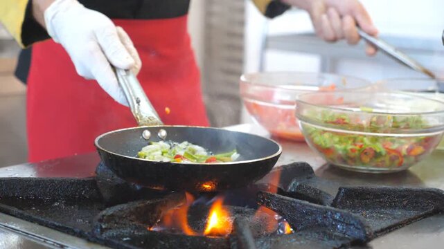 Unrecognizable chef holding two pans and cooking flambe style dish in modern kitchen of restaurant. Male cook mixing and frying vegetables in flaming skillets on stove. Concept of cooking. Slow motion