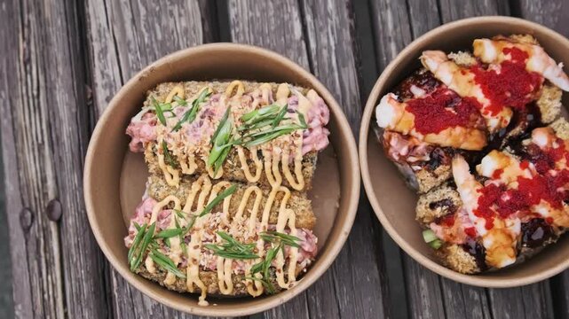 Overhead perspective shows two paper bowls containing fried sushi dogs and shrimp rolls. The flat lay composition contrasts the colorful food against the rustic wooden planks.