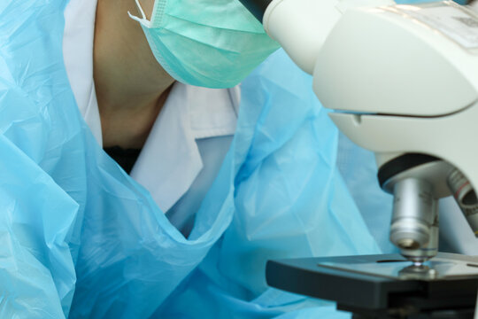 Laboratory scientist in protective coat and mask examines sample under microscope with sterile blue gown and focused posture