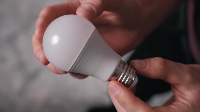 Close-up of male hands holding and rotating a white energy-efficient LED light bulb. The focus on the frosted dome and screw base illustrates modern illumination technology.