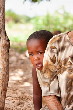 Young African Boy Peeking From Behind Mother Outdoors in Rural Village Settlement