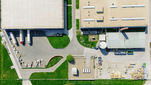 Aerial view of an industrial warehouse complex with trucks at loading docks, wooden pallets, and large flat-roofed buildings surrounded by green grass in Sremska Mitrovica, Vojvodina, Serbia.
