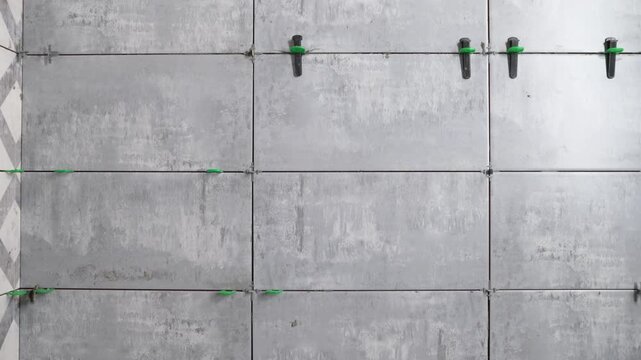 Closeup of a construction worker installing large gray ceramic tiles on a wall. Tiler uses a professional tile leveling system with green clips black wedges for a perfect finish