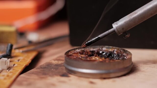 Extreme close-up slow motion of a hot soldering iron tip being dipped into a tin of smoking rosin flux. The violent bubbling reaction illustrates high heat and chemical cleaning.