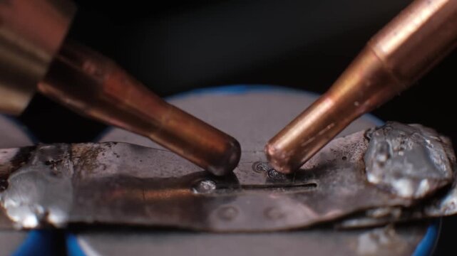 Macro slow motion shot of bright sparks flying as electrodes fuse a metal strip to a battery terminal. Dramatic lighting highlights the intensity of industrial resistance welding.