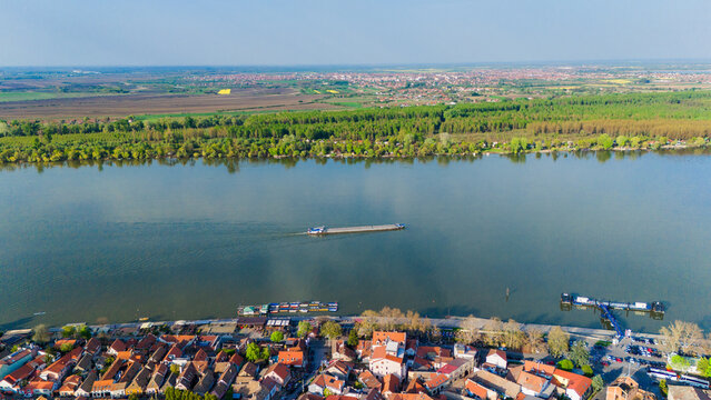Aerial view of the historic Zemun district and the Danube River with a cargo barge navigating the water near red-roofed houses and green riverbanks Belgrade, City of Belgrade, Serbia.