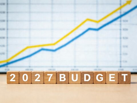 Wooden blocks with the text of the 2027 budget against a blurred stock market chart.