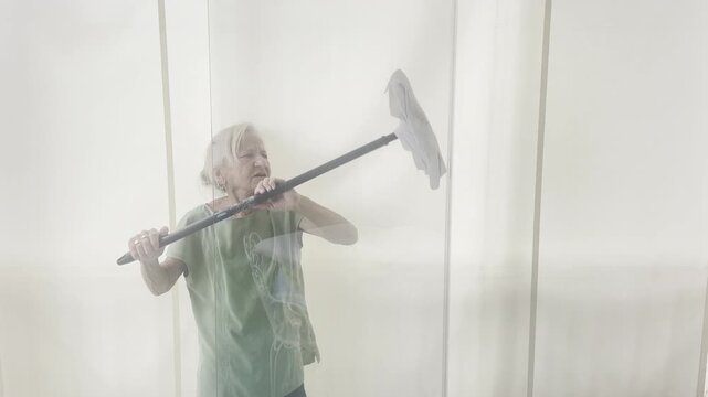 Elderly woman using a long-handled squeegee mop to clean a high glass window. Seen through the reflection, she works with effort and focus in a bright domestic setting