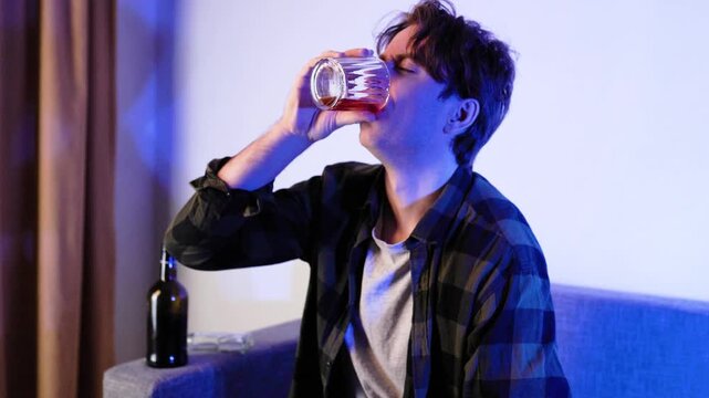 Sad depressed man drinking alcohol from a glass while sitting on a sofa. A bottle of alcohol stands nearby. Concept of alcoholism, addiction, divorce, unemployment, and loneliness. 4k footage