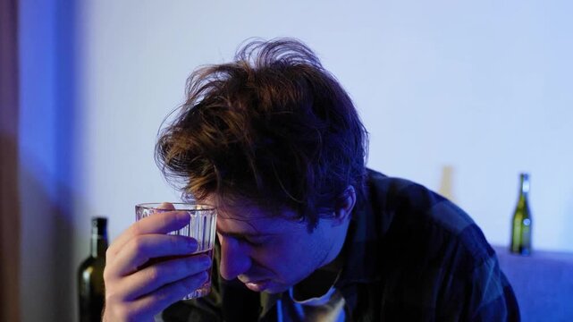 Close-up of a depressed, sad man holding a glass of alcohol to his forehead while sitting on a sofa. Concept of alcoholism, severe addiction, divorce, unemployment, and loneliness.4k footage