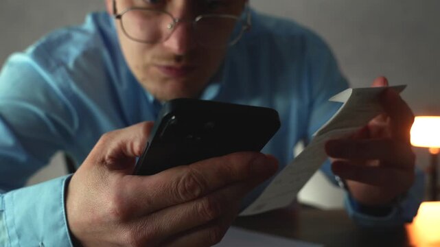 A man reviews a paper receipt while checking prices on his smartphone after noticing a possible billing discrepancy from a supplier invoice during office accounting.