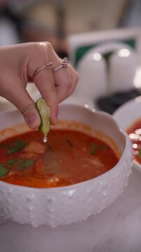 Female hand squeezing lime into tom yum soup in a bowl. Traditional Thai spicy soup served in a restaurant or at home