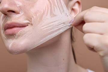 Fototapeta premium Beautiful woman peeling off facial mask on brown background, closeup
