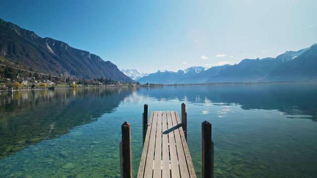 A serene perspective from a wooden pier extends into the crystal-clear turquoise waters of Lake Geneva with snow-capped Swiss Alps and a calm reflective surface stretching to the distant horizon.