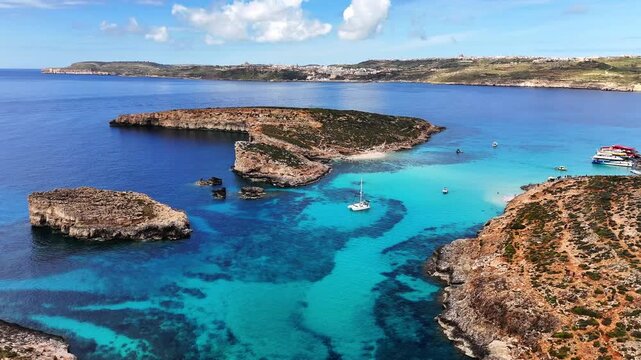 Blue Lagoon in Comino Għajnsielem Gozo Region Malta with shallow turquoise basin over carbonate seabed and limestone islets between Comino and Cominotto, clear Mediterranean water, slow drone pullback