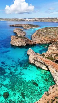 Blue Lagoon in Comino Għajnsielem Gozo Region Malta with limestone islets and shallow turquoise basin over carbonate seabed between Comino and Cominotto vertical orbit establishing shot
