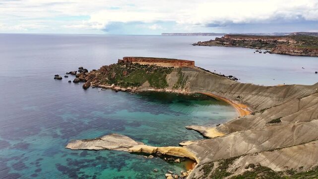 Qarraba Bay in Mġarr Malta with blue clay slopes beneath limestone plateau forming a curved cove over clear Mediterranean water within Ġnejna Bay system, drone push in and descend shot