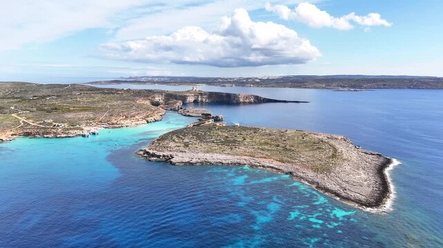 Blue Lagoon between Comino and Cominotto Kemmunett with Upper Coralline Limestone cliffs and St Mary Tower Torri ta Santa Marija overlooking Mediterranean Sea, high wide aerial shot