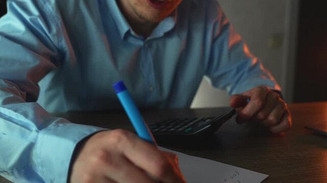 Businessman calculates financial data on a calculator while making handwritten notes to avoid mistakes, working late in a dim office. Concept of focus and accuracy under pressure.