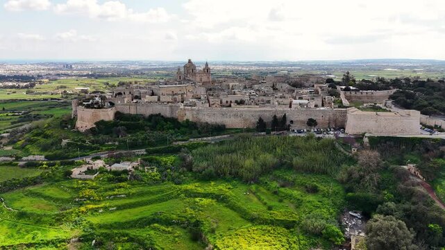 Mdina in Northern Region Malta viewed from south side showing fortified limestone bastions and St Paul Cathedral overlooking Rabat and agricultural valley with road network, orbiting aerial shot