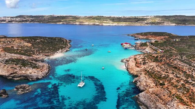 Blue Lagoon in Comino Għajnsielem Gozo Region Malta showing limestone shores with small landing jetty and anchorage zone with boats over shallow turquoise basin between Comino and Cominotto, aerial