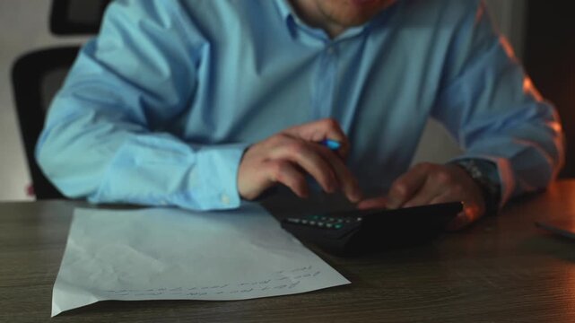 Businessman calculates financial data on a calculator while making handwritten notes to avoid mistakes, working late in a dim office. Concept of focus and accuracy under pressure.