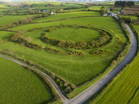 Aerial view of Castle Dore hillfort surrounded by lush green fields, trees, and winding country roads in Par, England, United Kingdom.