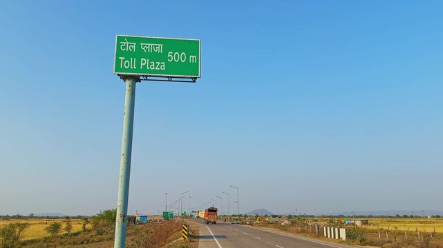 Wide highway scene featuring a green &ldquo;Toll Plaza 500 m&rdquo; sign in Hindi and English on a tall pole, with a heavy vehicle moving away in the background through open rural farmland under a clear blue sky.