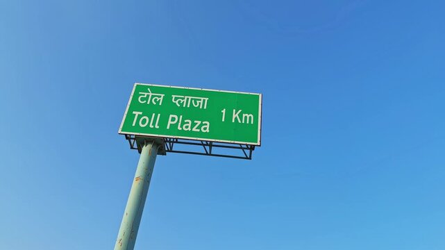 Low-angle view of a green highway sign reading &ldquo;Toll Plaza 1 Km&rdquo; in Hindi and English, mounted on a tall pole against a clear blue sky, captured with a subtle orbiting motion.