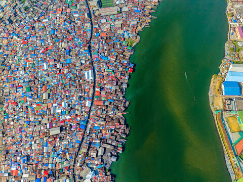 Aerial view of a dense patchwork of colorful rooftops sprawl beside the murky green river waters, a stark contrast to the industrial landscape, Monrovia, Liberia.