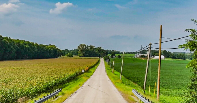 Aerial view of a long, straight road carving through vibrant golden corn and deep green fields towards distant farm buildings under a vast blue sky, Lebanon, Ohio, United States.