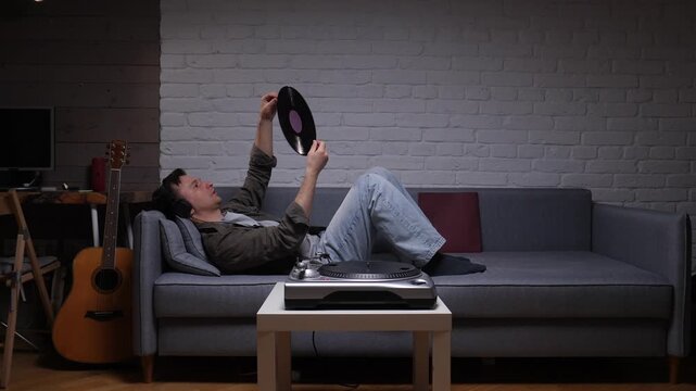 Man lying on a sofa with headphones, carefully holding a vinyl record above his head, ready to place it on the turntable for an evening of nostalgic music listening at home