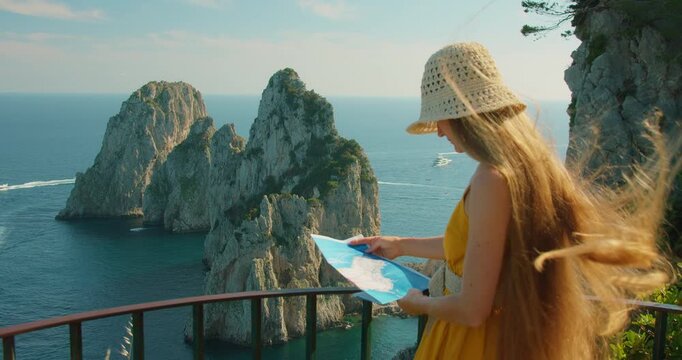 Curious woman comparing her paper map with the Faraglioni view in Capri on a summer day