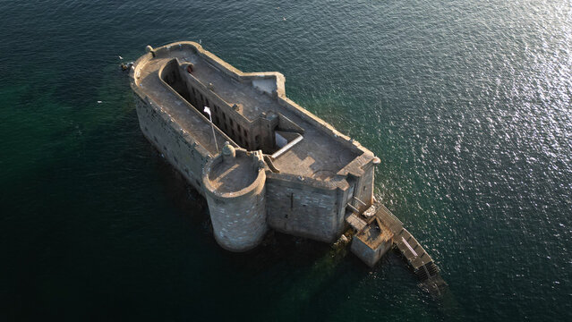 Aerial view of Chateau du Taureau, a stone fortress rising from the dark water, its stark silhouette contrasting with the shimmering surface, Plouezoc'h, Bretagne, France.