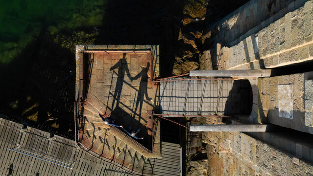 Aerial view of ethereal shadows of two figures holding hands stretching across rugged stone walls and a wooden pier, Plouezoc'h, Bretagne, France.