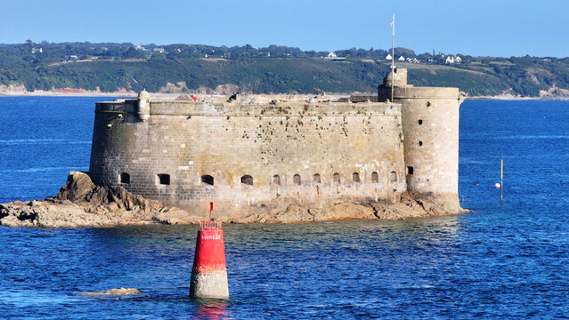 Aerial view of a weathered stone fort on a rocky island in deep blue waters, a red and white buoy, green coastline, Plouezoc'h, Bretagne, France.