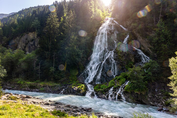 Mountain waterfall cascades rocky cliff fast river dense forest alpine landscape. Bright sunlight flare mist while clear water flows wilderness evoking fresh nature travel tranquil scenery © Kyryl Gorlov