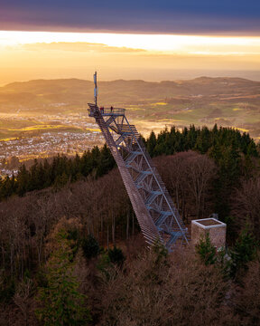 Aerial view of the striking Trommturm observation tower piercing a warm golden sky, overlooking a serene valley town amidst lush forest, Rimbach, Hessen, Germany.