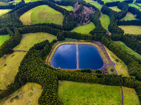 Aerial view of geometric patterns of dark green tree lines embracing bright emerald fields, with a serene deep blue reservoir at its heart, Terceira, Azores, Portugal.